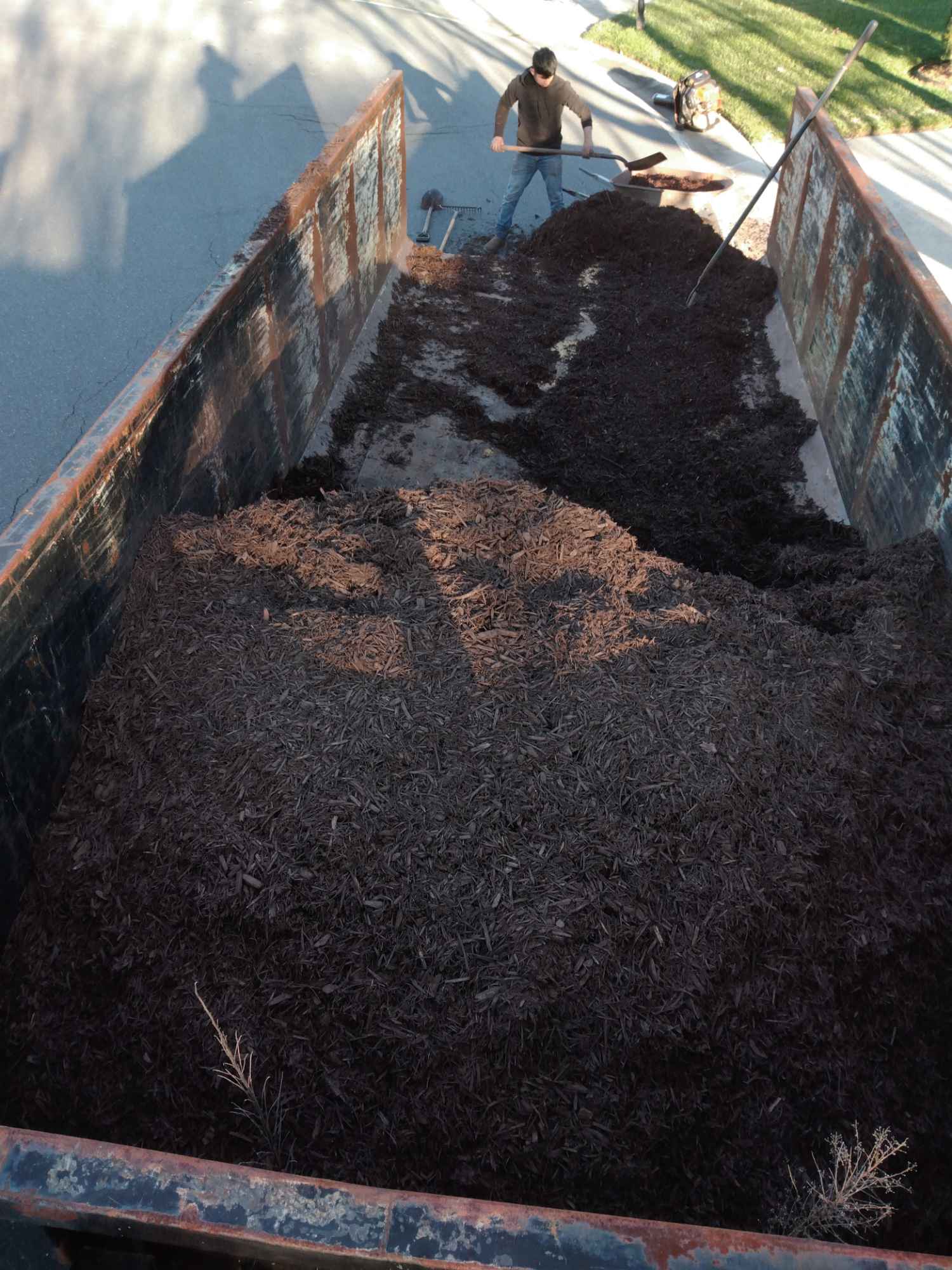 A person shoveling dark mulch from a large, rusted metal container on a sunny day, with nearby grass visible.