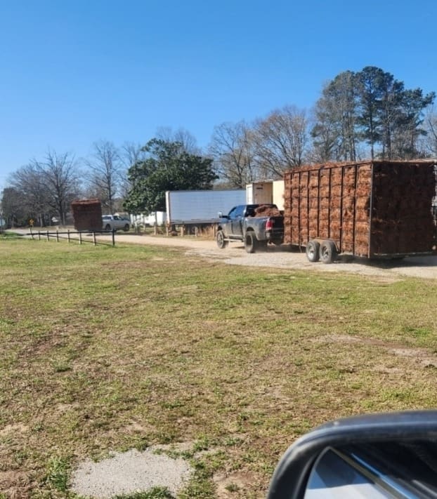 A pickup truck is hitched to a large, rust-colored trailer filled with baled pine straw
